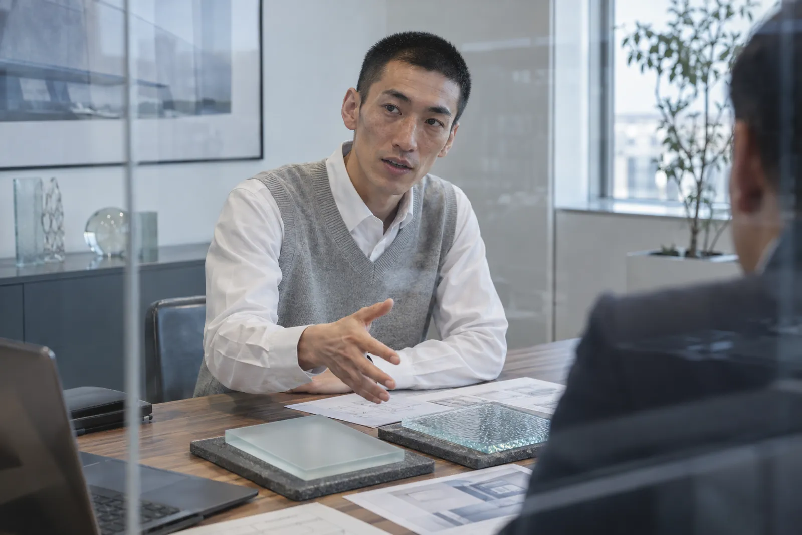 打ち合わせ室で図面とフロスト・型板のサンプルを示し、設計条件を説明する技術営業課の社員